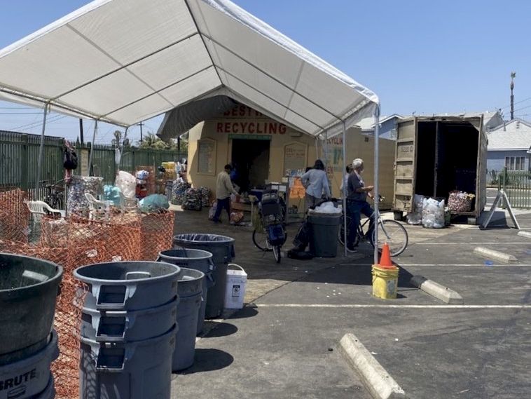 An outdoor recycling site with a large tent labeled &ldquo;Recycling,&rdquo; people sorting items, and many trash/recycling bins and bags.