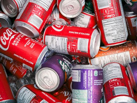 A pile of Coca‑Cola soda cans&mdash;mostly red and purple&mdash;showing &ldquo;Original Taste&rdquo; and &ldquo;Coke&rdquo; labels on metallic tops.